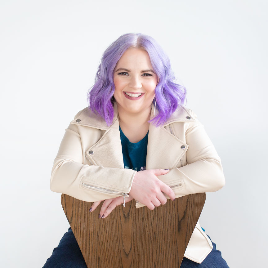Woman with colorful hair in a leather jacket seated on a chair facing forward.